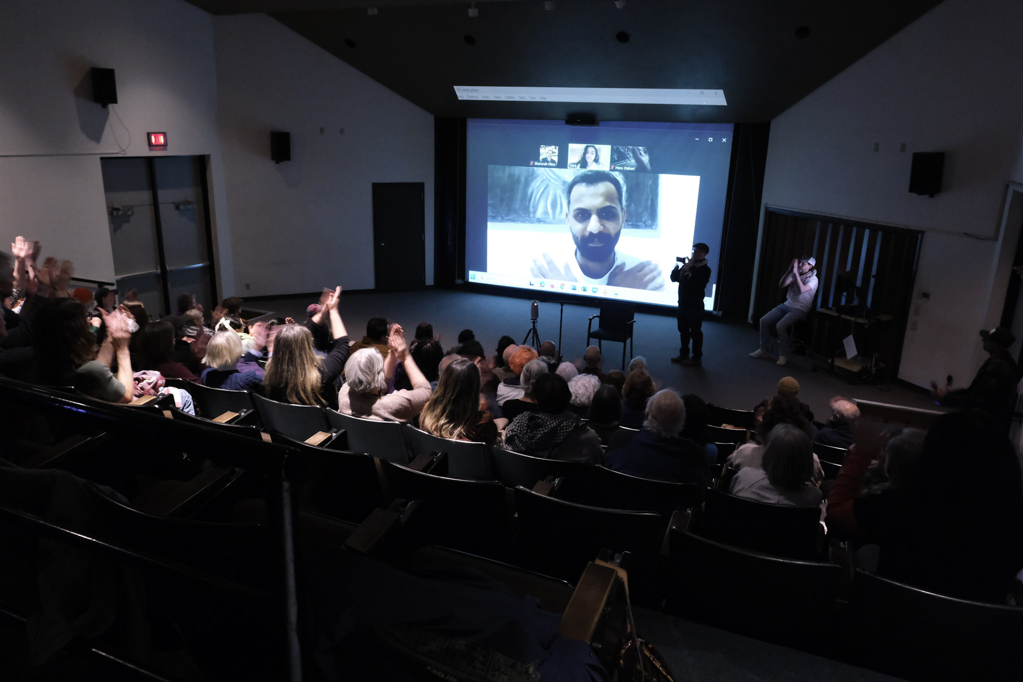 Screening audience watching Omar on a large projection screen during a live video call from Ramallah, with people clapping toward the screen.